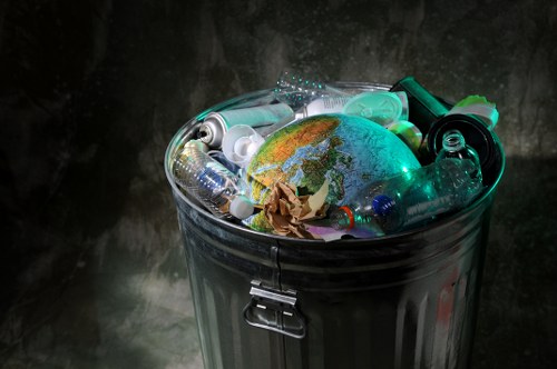 A cylindrical metal rubbish bin with a closed, shiny silver lid is filled with various types of waste, including clear plastic bottles, a crumpled piece of brown paper, and other small plastic containers. The bin is situated outdoors against a dark, textured background, possibly a weathered wall or surface. The waste visible includes transparent and coloured plastic items, and the bottles are of different shapes and sizes, some with caps and some without. The transparency and reflective qualities of the plastic objects contrast with the metallic finish of the bin. The scene is lit with soft, diffused lighting that highlights the textures of the plastic and the metallic surface, suggesting a typical rubbish collection environment. This image exemplifies typical waste disposal practices in urban areas, such as Hackney, and aligns with professional rubbish removal services like those offered by Commercial Waste Removal Hackney, specialising in the collection and handling of general waste materials.