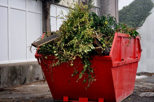 A large red plastic skip filled with a mixture of green garden waste, including leafy branches, shrub clippings, and small twigs, situated outdoors on a paved area next to a white-painted wall with a window. The skip's open top reveals the dense, leafy plant material inside, with some branches leaning over the edge. In the background, there is a concrete base and part of a brick wall, with a hedge visible further in the distance. The environment suggests a garden or back yard area where garden waste is being prepared for collection or disposal, consistent with rubbish removal services such as those offered by Commercial Waste Removal Hackney.