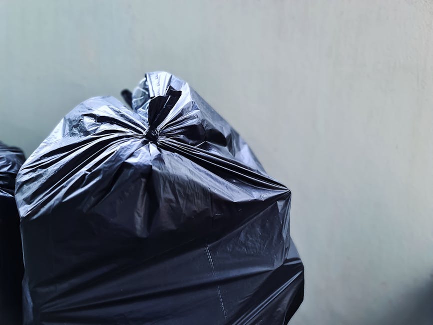 A large pile of transparent plastic rubbish bags filled with mixed waste, including clothing, paper, and packaging materials, situated on a paved sidewalk outside a commercial establishment at night. 